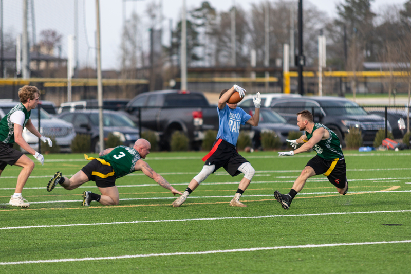 A flag football player in a blue jersey catches the ball mid-air as three opposing players in green jerseys attempt to intercept. Energetic and tense action.