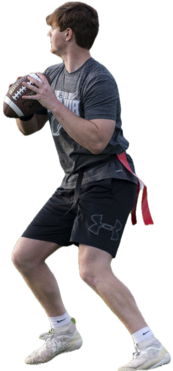 A man in athletic gear poised to throw a football, wearing a gray T-shirt, black shorts, and white cleats. A red flag belt suggests a flag football game.