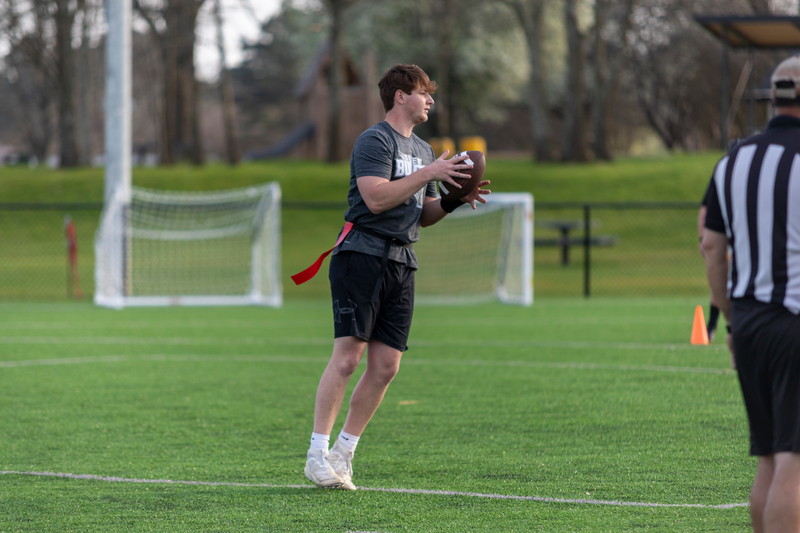 A young man prepares to throw a football on a grassy field, wearing a gray shirt and black shorts. A referee stands nearby. The mood is focused and athletic.