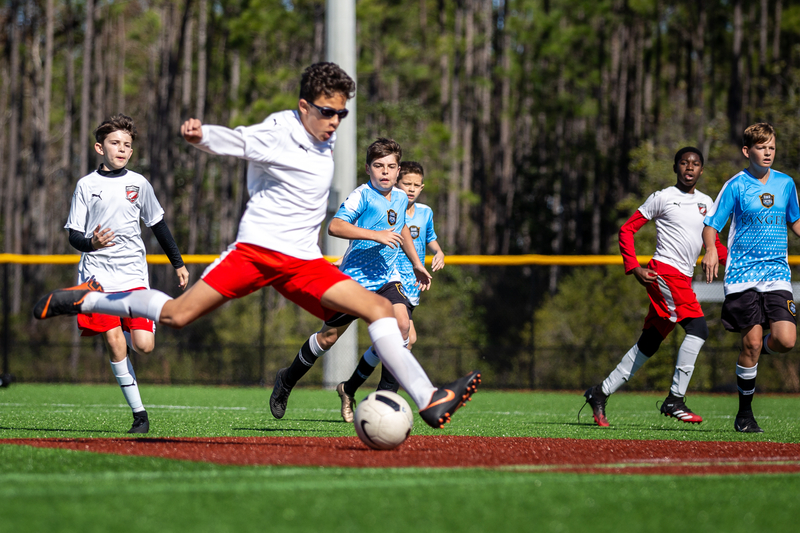A youth soccer game on a clear day. A player in a white shirt and red shorts kicks the ball mid-air. Opponents in blue jerseys and teammates follow.