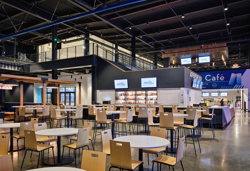 Modern cafe with light wood tables and chairs, high ceilings, and a sleek bar area. Signs read "Adventure Cafe," conveying a contemporary and inviting tone.