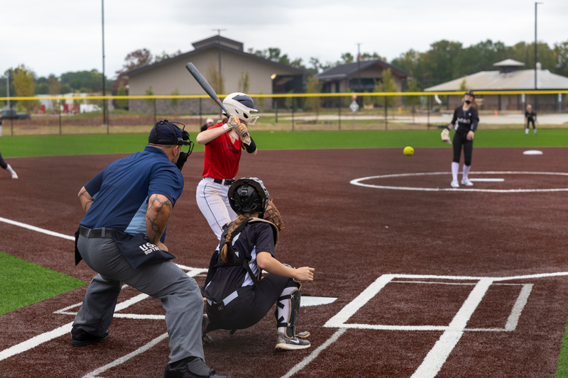 A softball game with a pitcher throwing to a batter in red. The catcher and umpire are positioned behind the batter. The field is bordered by trees.