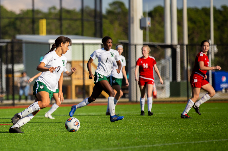 A girls' soccer game in action. A player in a white jersey dribbles the ball while teammates and opponents in red run on a sunny field.