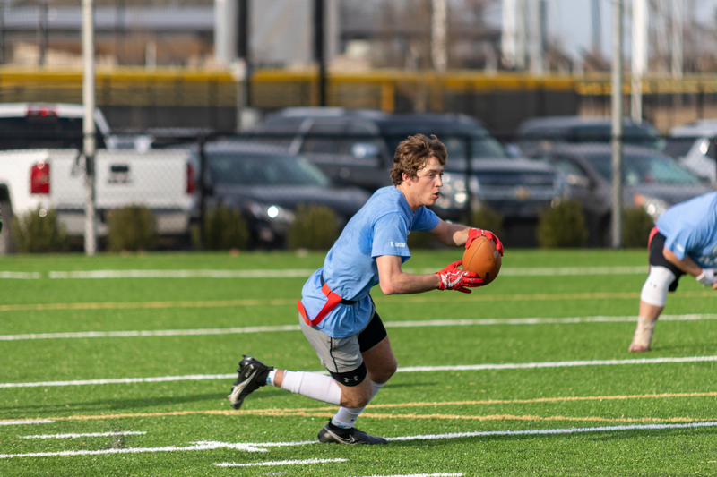 A young man in a blue shirt and red flag shorts runs on a green field, holding a football. He appears focused, with cars parked in the background.