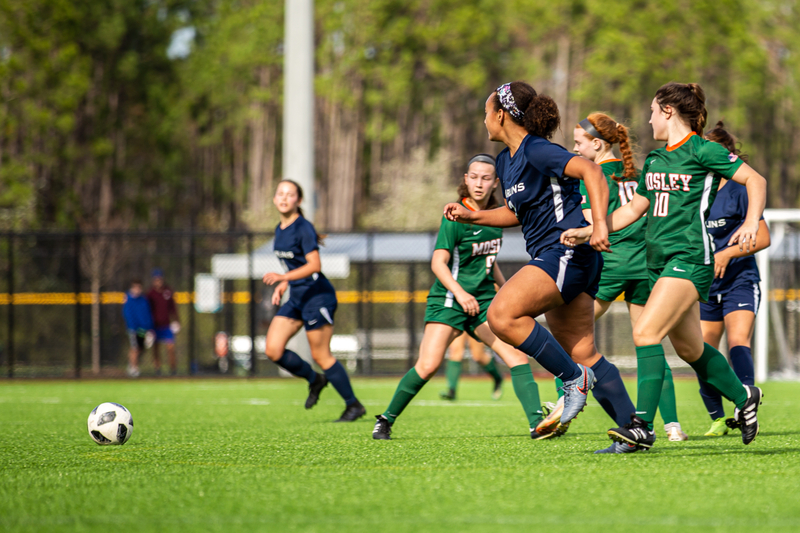 A group of female soccer players in green and blue jerseys chase a soccer ball on a lush green field, conveying action and teamwork in a sunny setting.